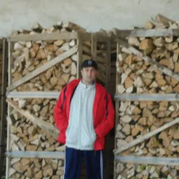 Mężczyzna w czerwonej kurtce stoi na tle drewnianych skrzyń wypełnionych pociętym drewnem opałowym.