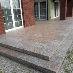 Zewnętrzny taras domu z dwoma stopniami wykonanymi z szarej, gruboziarnistej nawierzchni. Na pierwszym planie widoczna kostka brukowa i żółte kwiaty. W tle fragment elewacji z czerwonej cegły...