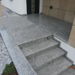 Zewnętrzne schody i taras wyłożone szarym, ziarnistym kamieniem, prowadzące do drzwi wejściowych, z dekoracyjnym żwirem i zielenią po bokach.