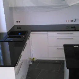 Nowoczesna kuchnia z czarnym blatem, białymi szafkami i czarną podłogą; widoczne przygotowane otwory na gniazdka elektryczne i folia ochronna na szafkach górnych.