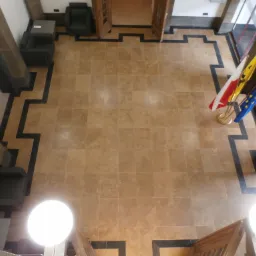 Widok z góry na hol wejściowy w Trąbkach Małych z beżową, kamienną posadzką ułożoną w kwadraty, ciemne obramowanie, flagi Polski i Unii Europejskiej oraz ciemne fotele.