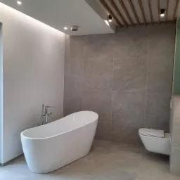 Nowoczesna łazienka z wolnostojącą białą wanną, podwieszaną toaletą i szarymi płytkami imitującymi kamień, oświetlenie punktowe i listwa LED, drewniane lamele na suficie.