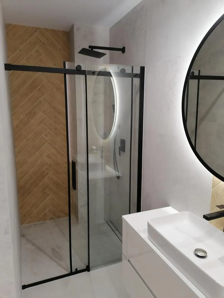 Nowoczesna łazienka z kabiną prysznicową z czarnymi ramami, deszczownicą, owalnym lustrem z podświetleniem LED i umywalką nablatową na białej szafce.