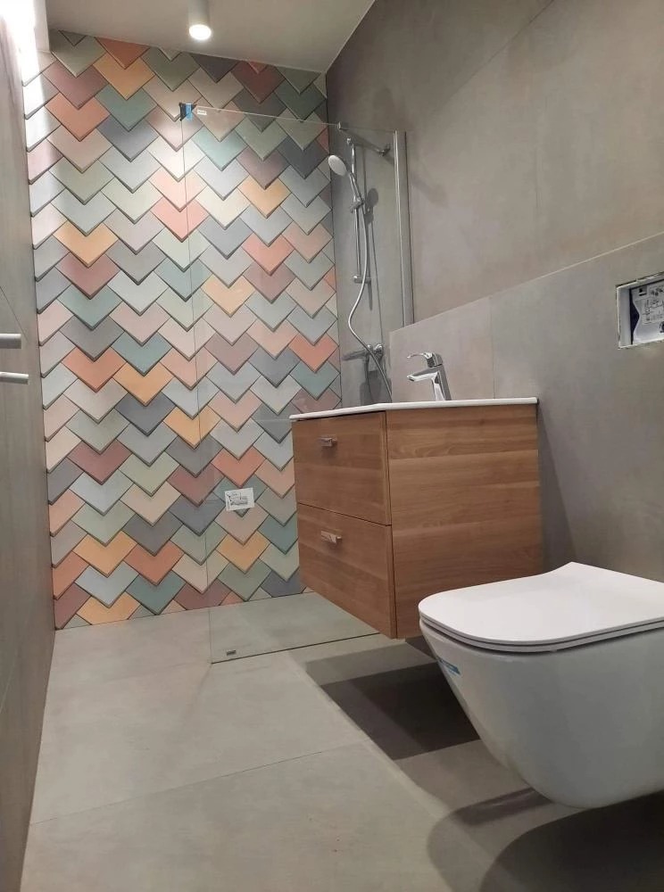 Wykończona łazienka z prysznicem typu walk-in, umywalką z szafką z jasnego drewna i podwieszaną toaletą; ściana prysznicowa wyłożona dekoracyjnymi, wielokolorowymi płytkami w kształcie strzałek.