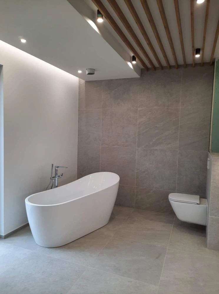 Nowoczesna łazienka z wolnostojącą białą wanną, podwieszaną toaletą i szarymi płytkami imitującymi kamień, oświetlenie punktowe i listwa LED, drewniane lamele na suficie.