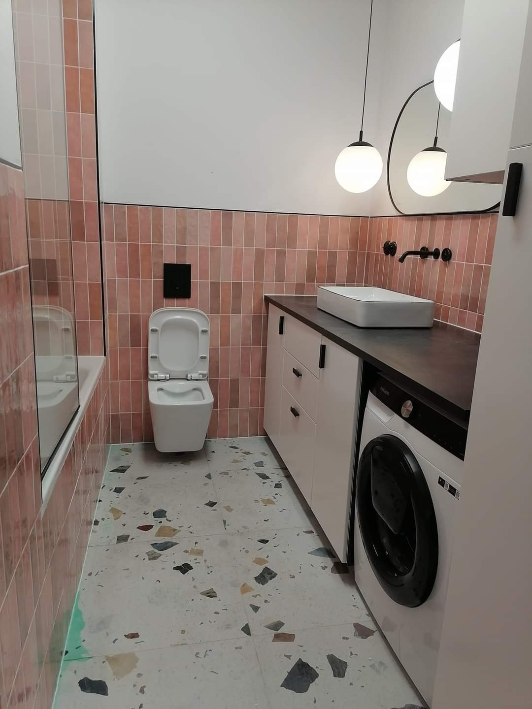 Nowoczesna łazienka po remoncie: różowe płytki typu cegiełka, biała toaleta podwieszana, umywalka nablatowa, pralka w zabudowie, podłoga lastryko i okrągłe lampy wiszące.