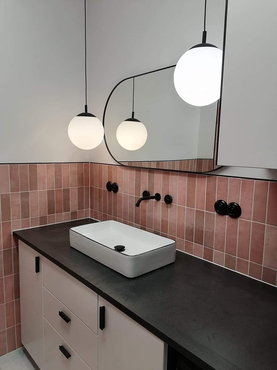 Łazienka z umywalką nablatową, różowe płytki na ścianie, czarna armatura i okrągłe lampy wiszące, minimalistyczny design.