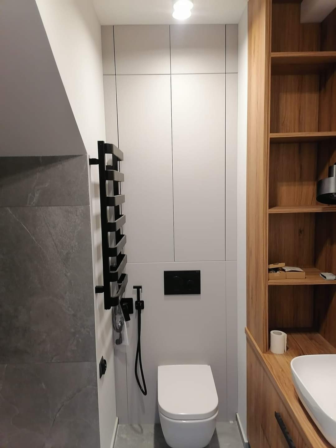 Nowoczesna łazienka z wiszącą toaletą, czarnym grzejnikiem drabinkowym i zabudową meblową z drewnianymi półkami oraz szarymi frontami.