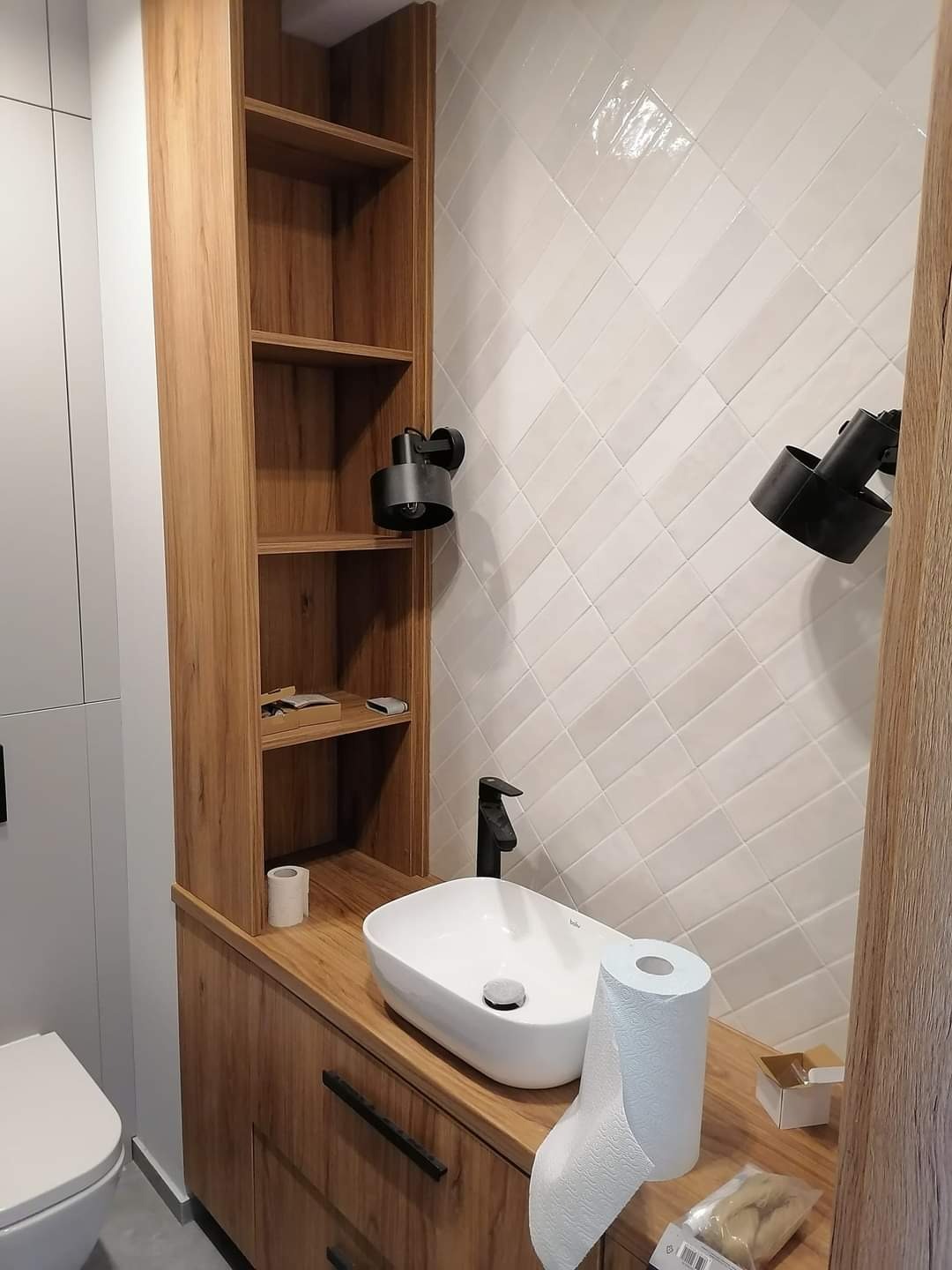 Nowoczesna łazienka z drewnianą szafką z półkami, białą umywalką nablatową i czarną armaturą, oświetlona dwoma punktowymi lampami na białej ścianie z ukośnych płytek.