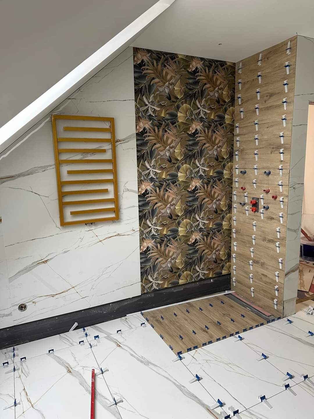 Łazienka w trakcie remontu: białe marmurowe płytki na ścianie i podłodze, złoty grzejnik drabinkowy, ściana z tapetą w motywy roślinne, fragment ściany wyłożony płytkami imitującymi drewno...