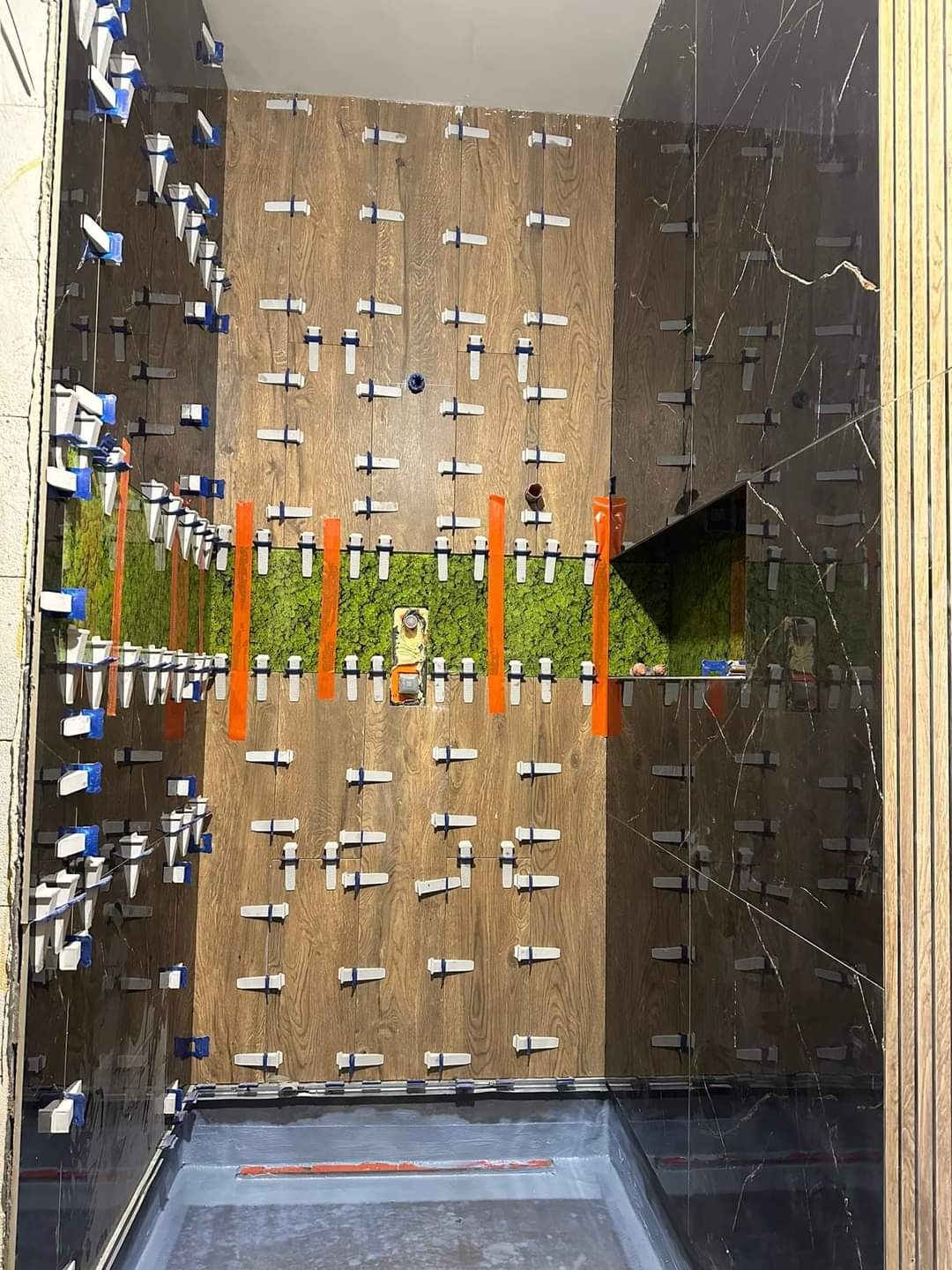 Montaż płytek imitujących drewno na ścianie łazienki z użyciem klipsów poziomujących, widoczny fragment zielonej ściany z mchu i czarne płytki podłogowe.