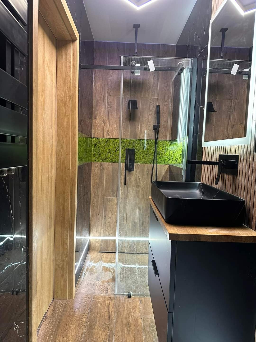 Nowoczesna łazienka z kabiną prysznicową typu walk-in, wykończona drewnopodobnymi płytkami i akcentem z mchu na ścianie, czarna umywalka nablatowa i armatura.