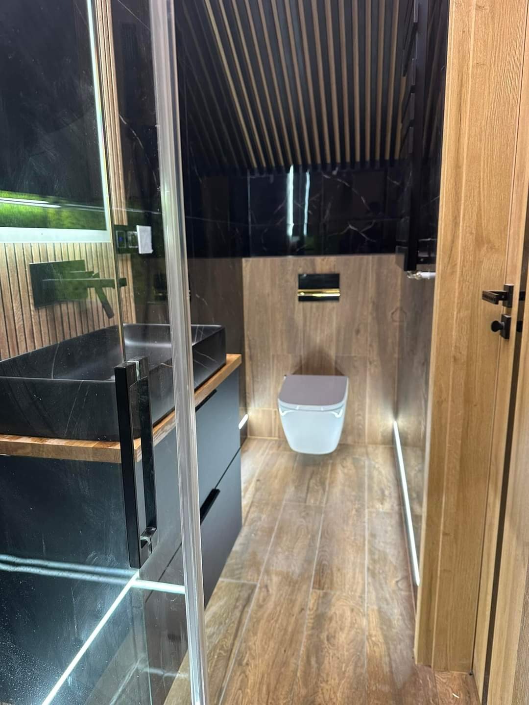 Nowoczesna łazienka z czarną umywalką nablatową, kabiną prysznicową ze szkła, drewnopodobnymi płytkami podłogowymi z podświetleniem LED i toaletą w odcieniach szarości.