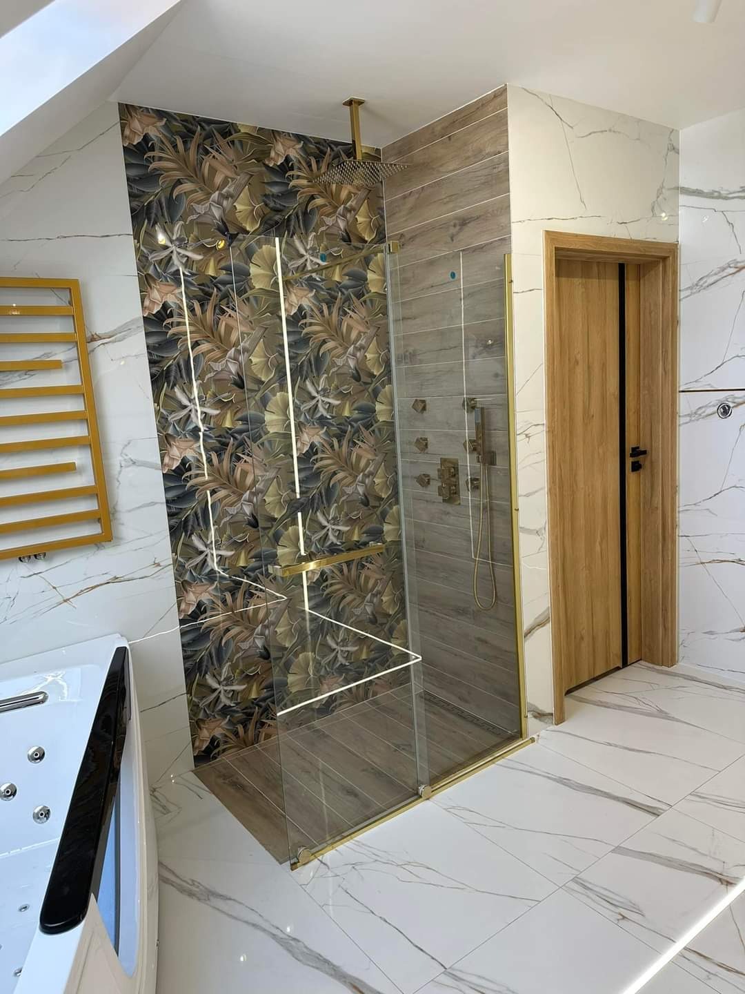 Nowoczesna łazienka na poddaszu z kabiną prysznicową typu walk-in, wykończoną złotymi detalami, z płytkami imitującymi drewno i wzorzystą tapetą z motywem roślinnym, oraz wanną z hydromasażem.