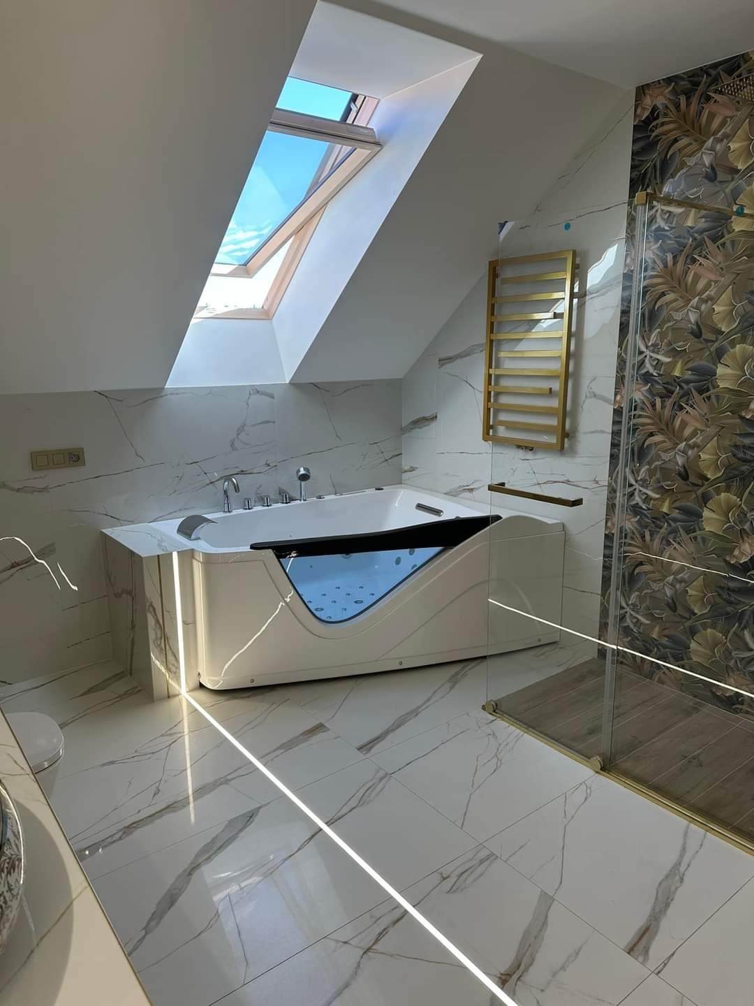 Nowoczesna łazienka na poddaszu z białą wanną z hydromasażem, marmurowymi płytkami i złotym grzejnikiem drabinkowym.