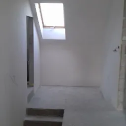 Poddasze w trakcie malowania, widoczne surowe schody, okno dachowe i wiszące kable elektryczne.