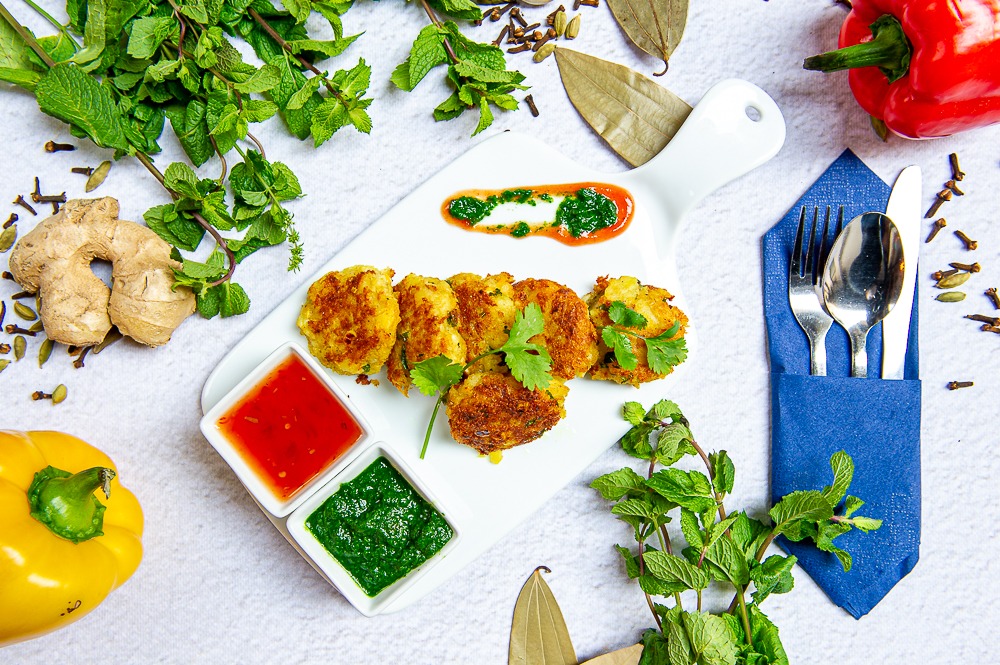 Elegancko podane kotleciki ziemniaczane na białej desce z uchwytem, z dwoma sosami w kwadratowych miseczkach, dekorowane świeżą miętą i kolendrą, obok papryka żółta i czerwona, imbir, sztućce...