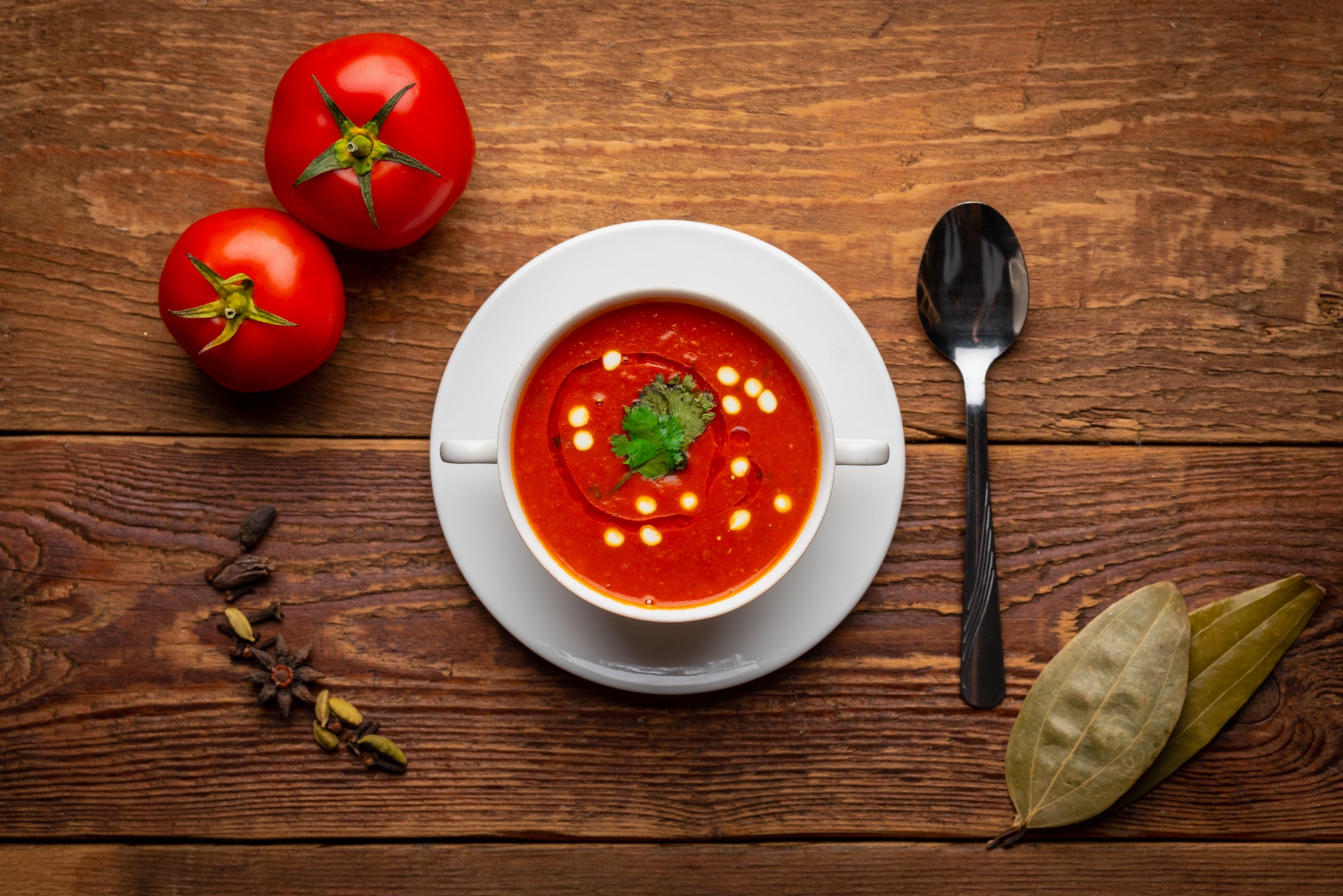 Krem pomidorowy w białej filiżance na talerzyku, udekorowany kleksem śmietany i listkiem kolendry, obok dwa pomidory, przyprawy i łyżka na drewnianym stole.