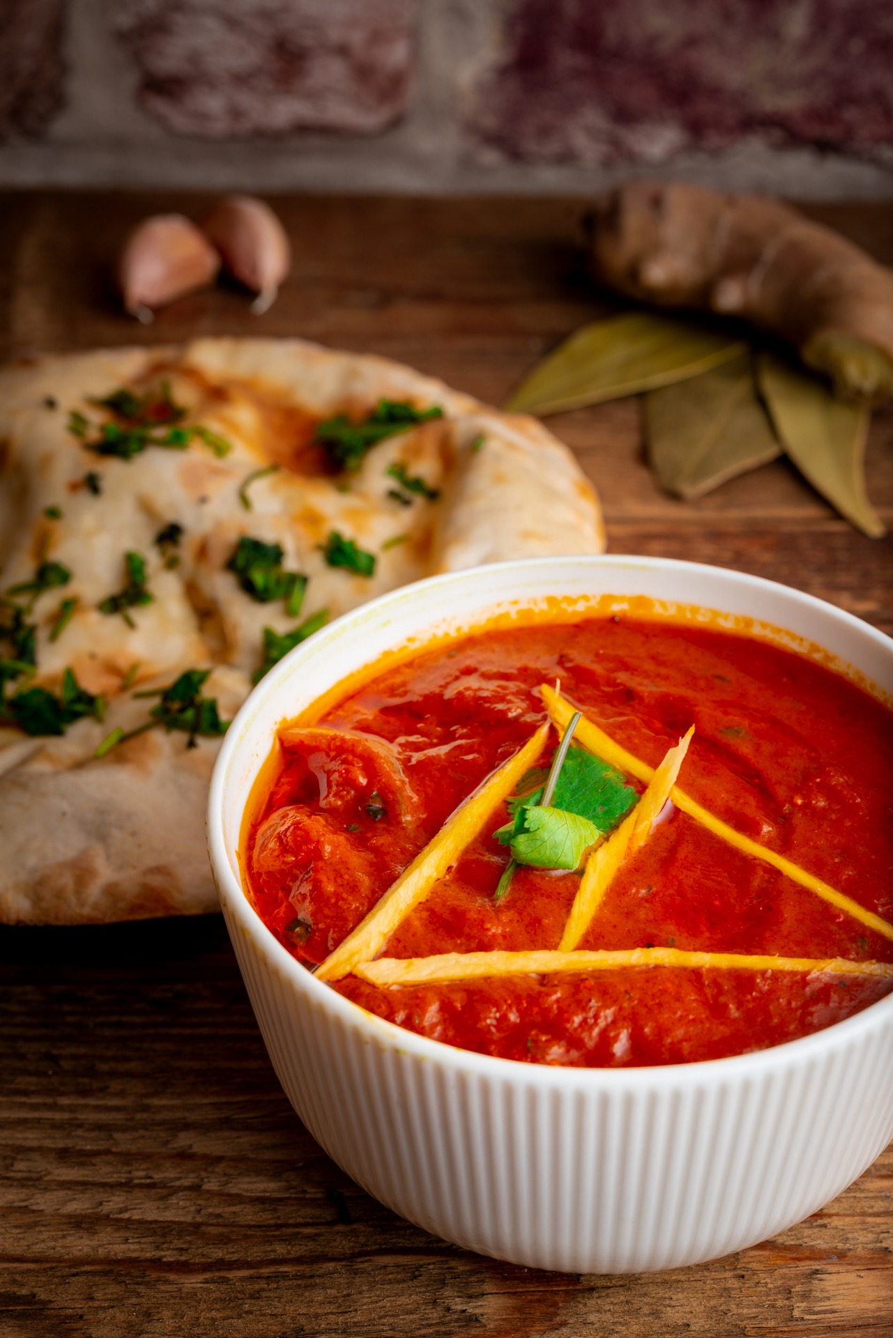 Indyjskie danie Butter Chicken w białej miseczce, udekorowane paskami i listkiem kolendry, obok chlebek naan posypany ziołami, czosnek i imbir na drewnianym tle.