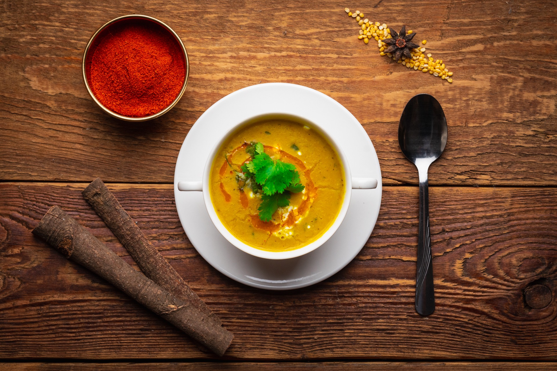 Kremowa zupa dyniowa z kolendrą i kroplami oleju, podana w białej miseczce na drewnianym stole z przyprawami i łyżką.