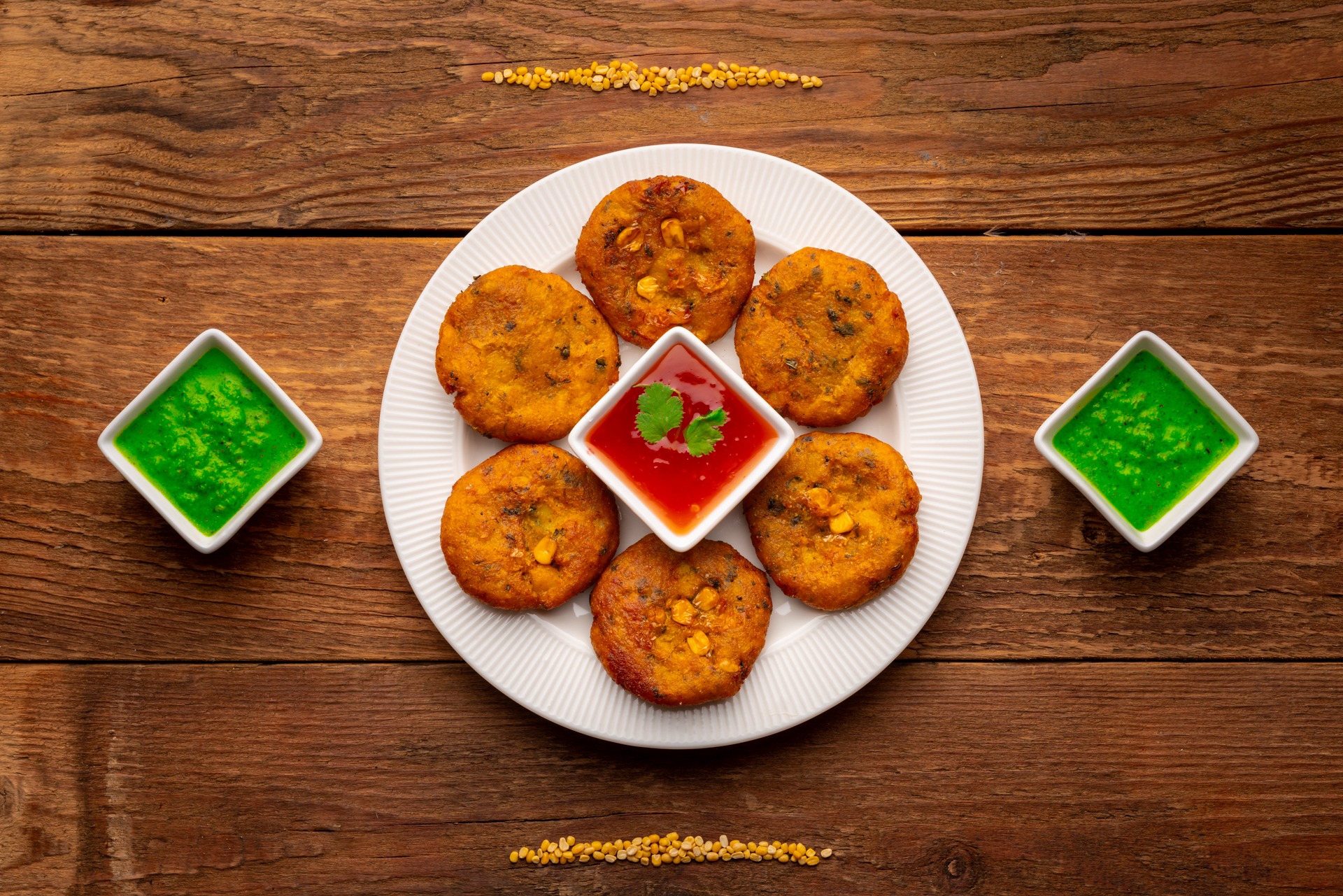 Smażone kotleciki z kukurydzą na białym talerzu, podane z sosem pomidorowym i zielonym sosem miętowym, ułożone na rustykalnym drewnianym stole z rozsypanymi ziarnami.