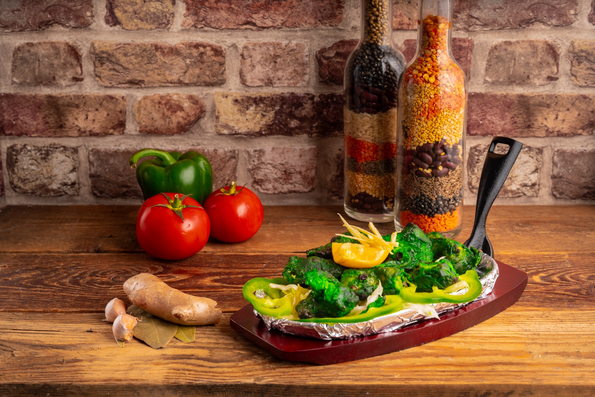 Zielone warzywa w sosie na aluminiowej folii, podane na drewnianej desce, obok imbir, czosnek, przyprawy w butelkach i czerwone oraz zielone papryki w tle