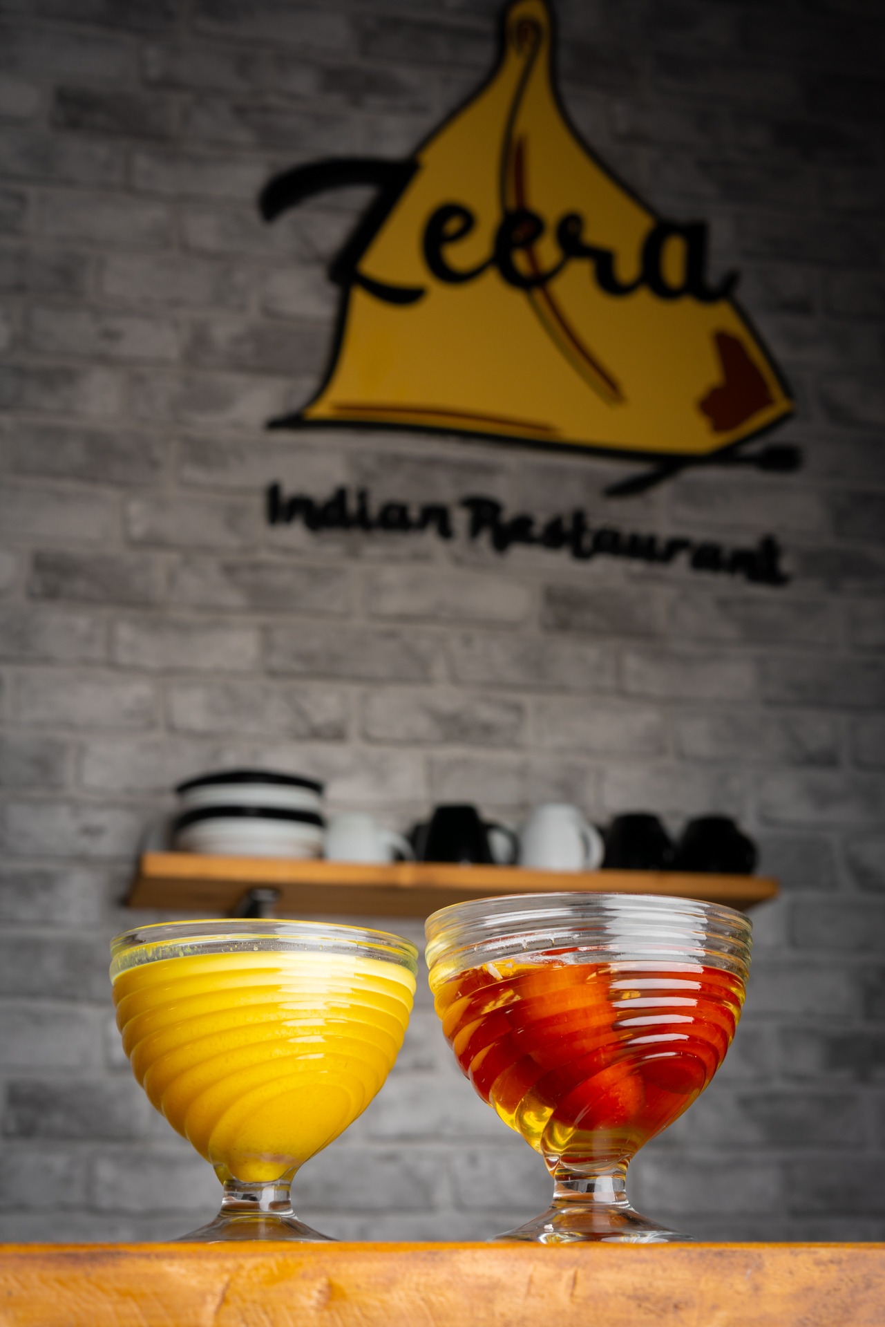 Dwa desery w szklankach na nóżkach: jeden z żółtym kremem, drugi z czerwonymi plasterkami owoców w syropie, na drewnianym blacie, w tle logo indyjskiej restauracji na tle ceglanej ściany.
