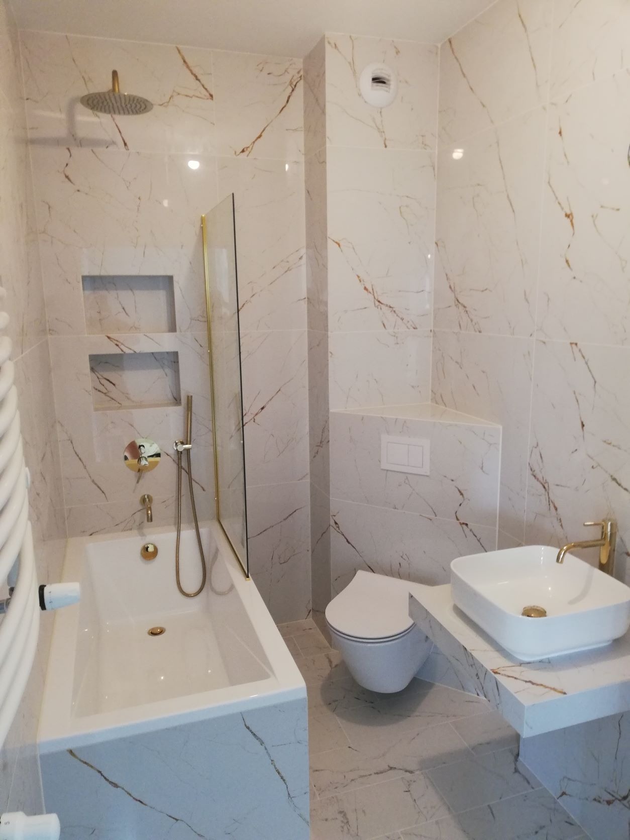 Wykończona łazienka z białymi płytkami imitującymi marmur z brązowymi żyłami, złota armatura, biała wanna i umywalka nablatowa.