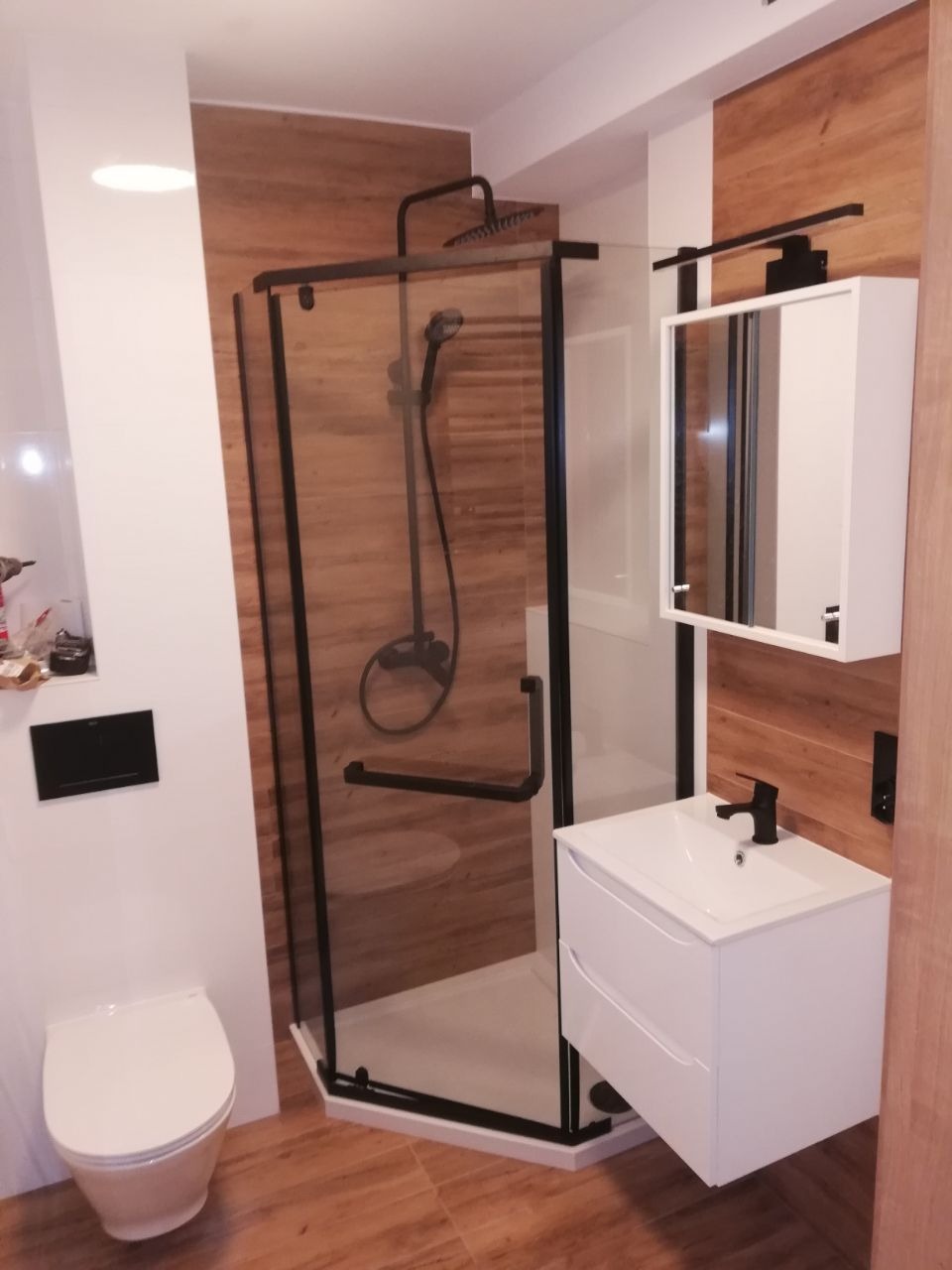 Nowoczesna łazienka z kabiną prysznicową typu walk-in z czarnymi profilami, białą umywalką z szafką i lustrem, oraz toaletą, wykończona płytkami imitującymi drewno na podłodze i ścianie.