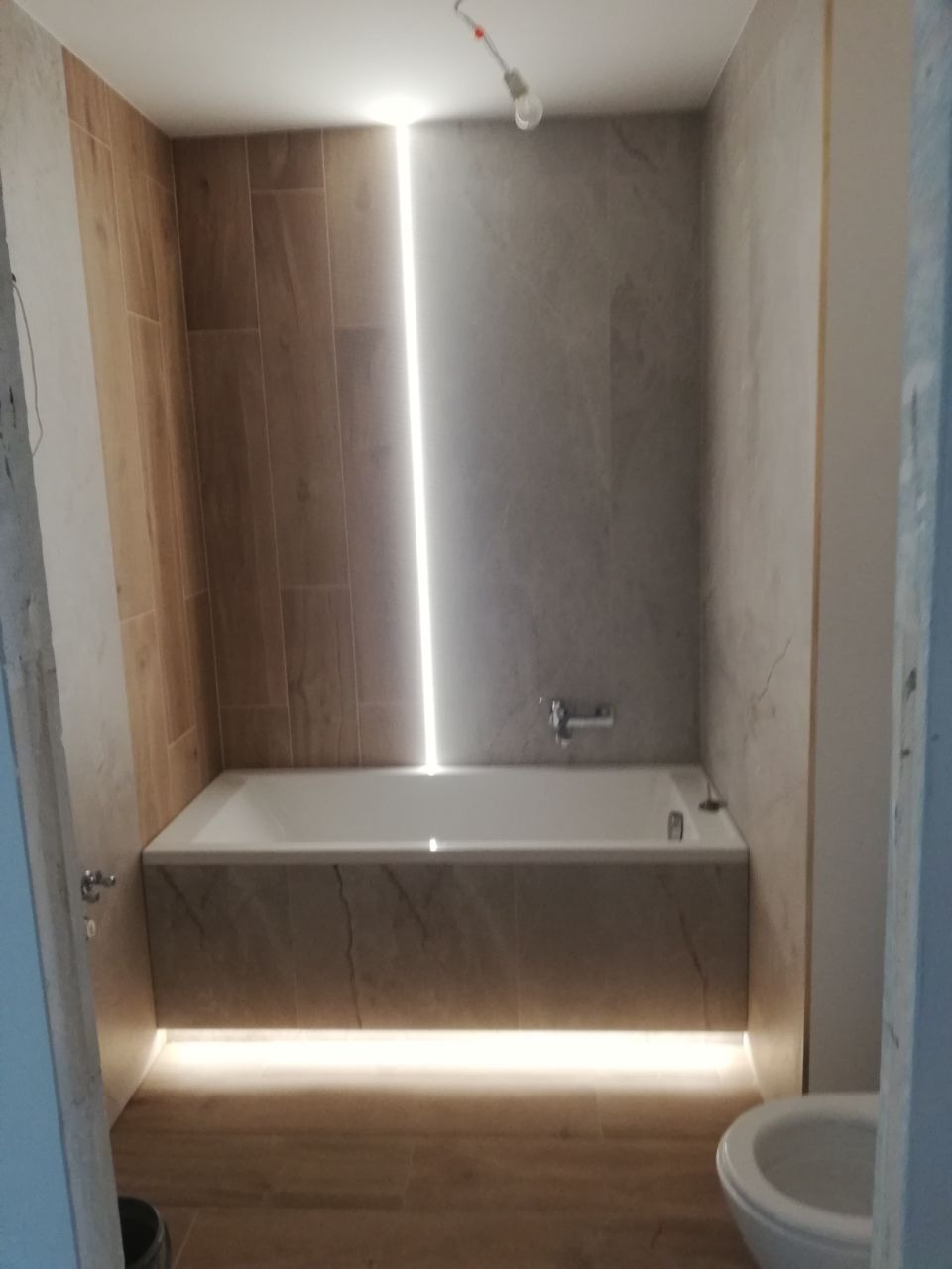 Łazienka z podświetlaną wanną, płytki imitujące drewno na ścianie i podłodze, pionowa listwa LED na ścianie, surowa żarówka zwisająca z sufitu.