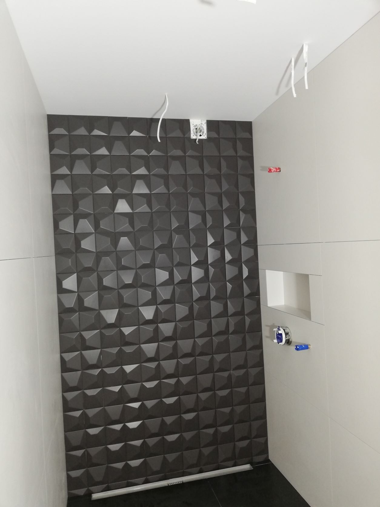Ściana w łazience wyłożona ciemnymi, trójwymiarowymi płytkami ceramicznymi z widocznymi wystającymi przewodami elektrycznymi i elementami instalacji wodnej.