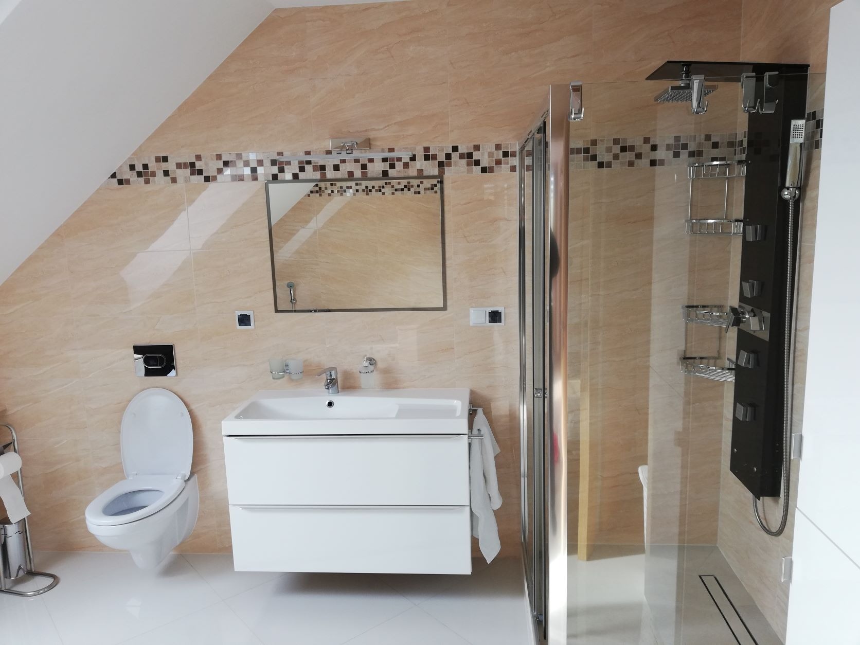 Jasna łazienka na poddaszu z beżowymi płytkami, mozaikowym pasem dekoracyjnym, białą umywalką z szafką, toaletą podwieszaną i kabiną prysznicową z panelem natryskowym.
