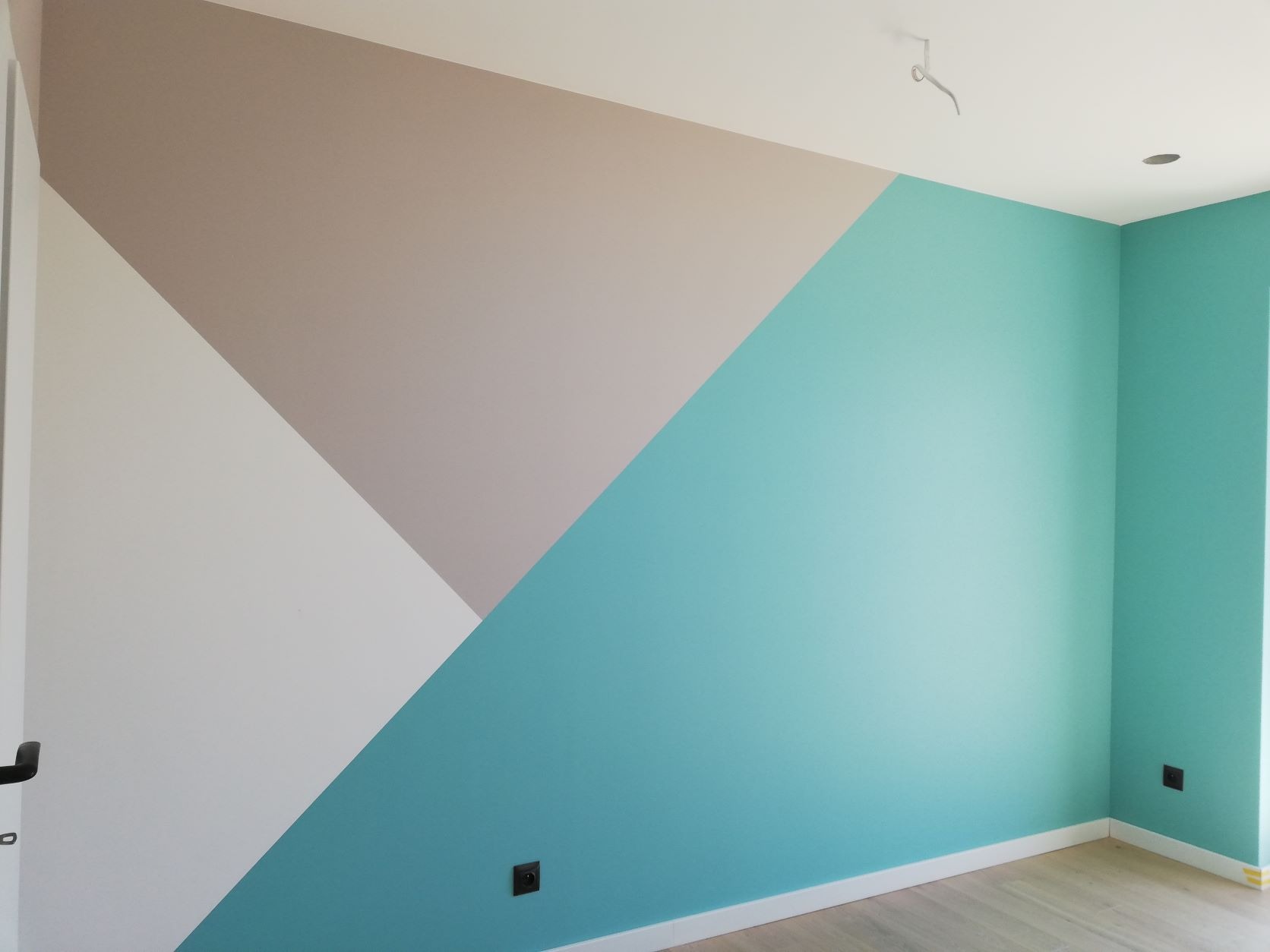 Ściana pokoju z geometrycznym wzorem malarskim w odcieniach bieli, beżu i turkusu; widoczne gniazdka elektryczne i listwy przypodłogowe.