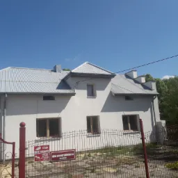 Dwupiętrowy budynek z jasną elewacją i metalowym dachem, widoczny zza czerwonego ogrodzenia, z tabliczkami informacyjnymi o monitoringu terenu i adresie.