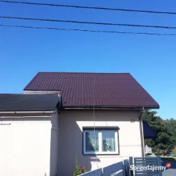 Dom jednorodzinny z nowym, brązowym dachem z blachodachówki, widok z dołu na tle błękitnego nieba. Na ścianie budynku widoczny numer 28.