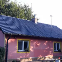 Dom jednorodzinny z ciemnym dachem z blachy falistej, różową elewacją i oknami z żółtymi ramami. Widoczny komin z czerwonej cegły i antena na dachu.