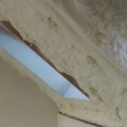 Poddasze z widoczną konstrukcją i zaaplikowaną pianką izolacyjną, tworzącą nierówną powierzchnię wokół otworu.