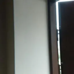 Wewnętrzny narożnik ściany pomalowanej na biało, z fragmentem ciemnobrązowej ościeżnicy okiennej i widocznym światłem dziennym.