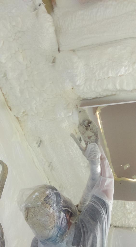 Pracownik w kombinezonie ochronnym aplikuje piankę poliuretanową na poddasze, widoczne nierówności i struktura pianki, metalowe elementy konstrukcyjne.