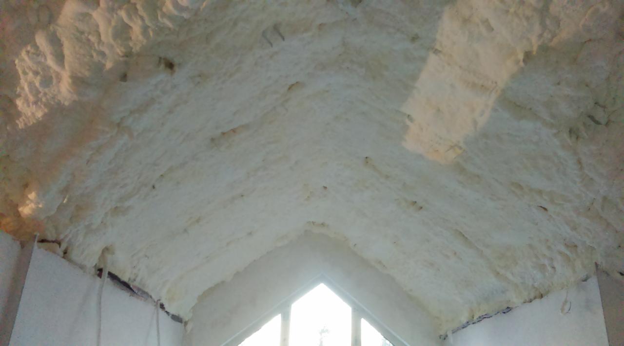 Poddasze ocieplone pianką poliuretanową, widok na skośny sufit z widocznymi nierównościami struktury pianki i fragment okna szczytowego w tle.