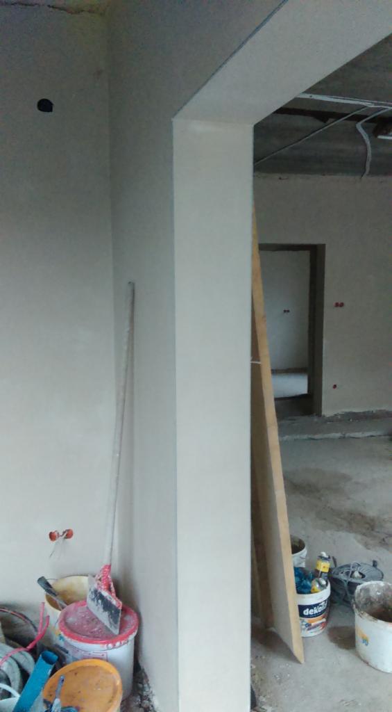 Wnętrze w trakcie prac wykończeniowych, widoczne świeżo otynkowane ściany, sufit w stanie surowym, narzędzia malarskie i wiadro z farbą przy ścianie.