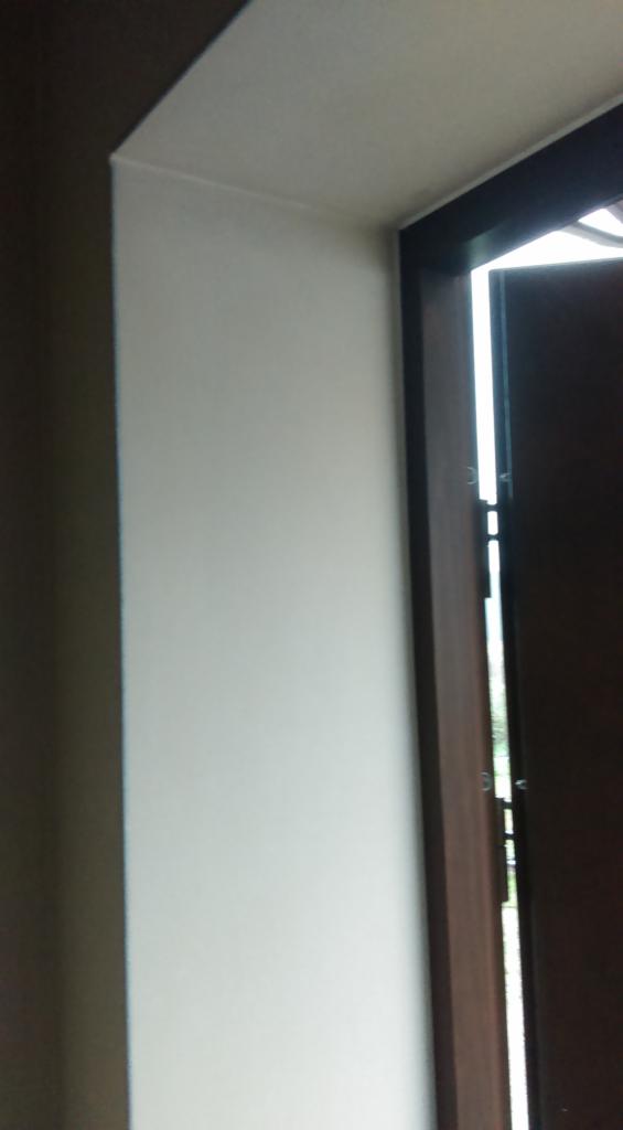 Wewnętrzny narożnik ściany pomalowanej na biało, z fragmentem ciemnobrązowej ościeżnicy okiennej i widocznym światłem dziennym.