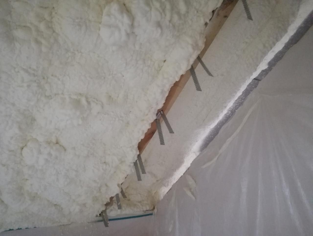 Poddasze z widoczną drewnianą konstrukcją i nałożoną, nierównomierną warstwą białej pianki izolacyjnej, zabezpieczone folią.