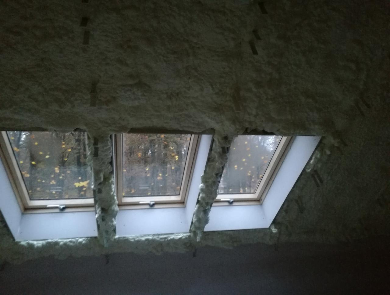 Poddasze z trzema oknami dachowymi po aplikacji pianki izolacyjnej PUR, widoczne fragmenty łat i drzewa z opadłymi liśćmi za oknami.