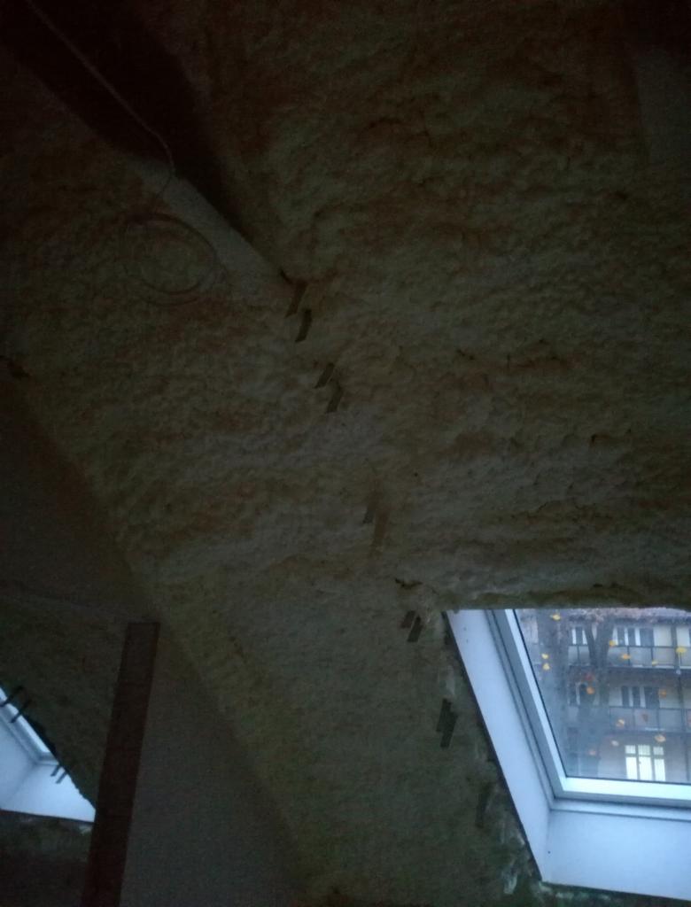 Poddasze ocieplone szarą pianką poliuretanową, widoczne okno dachowe i fragment konstrukcji drewnianej.