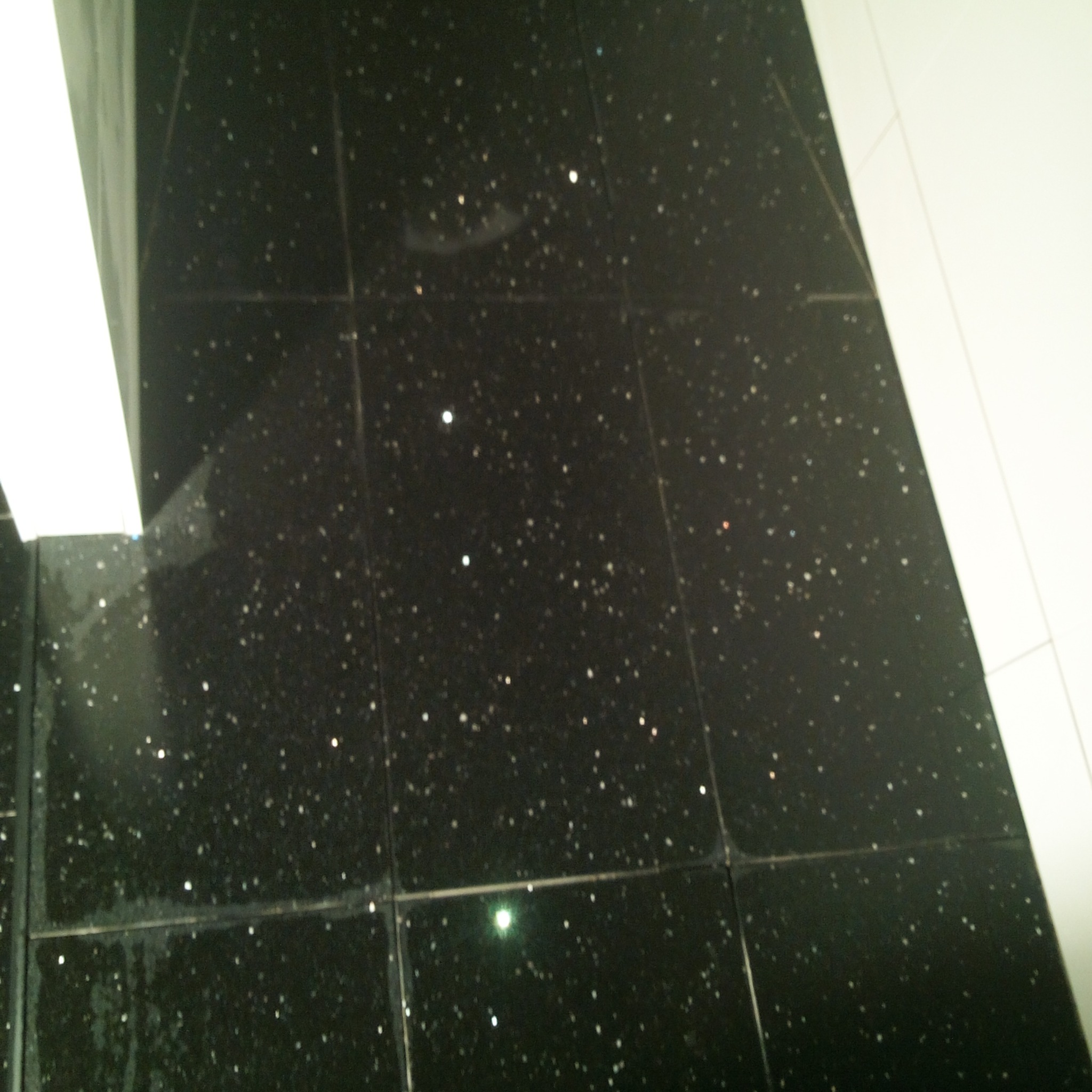 Czarna, błyszcząca podłoga wyłożona płytkami z brokatowym efektem, widoczne białe elementy armatury w tle.