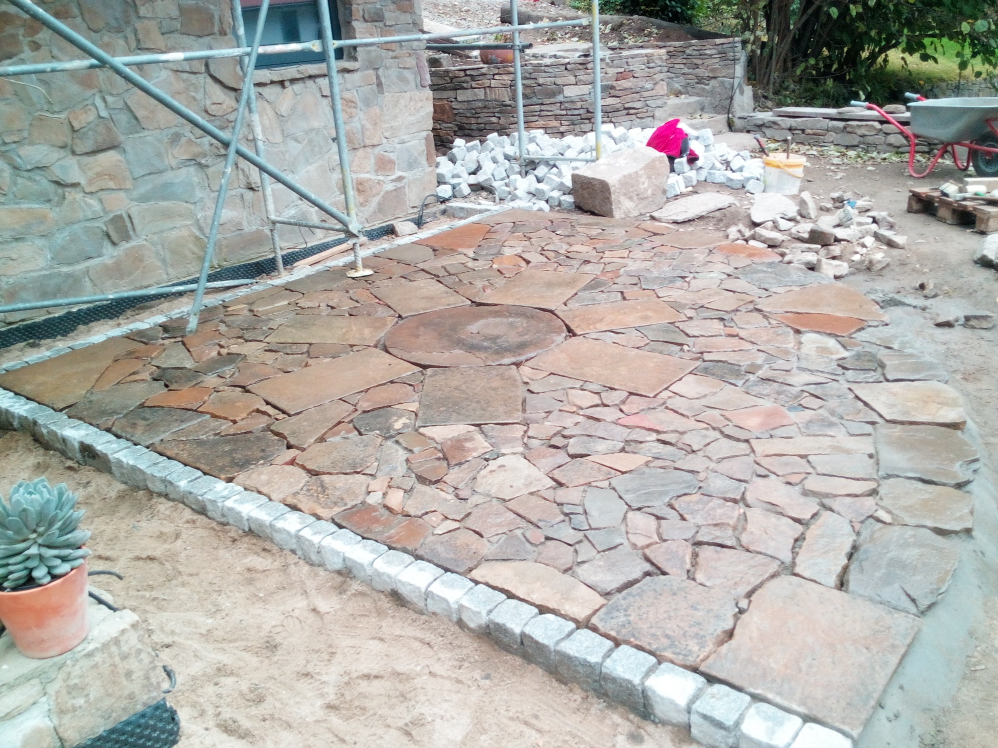 Układanie nieregularnej kamiennej nawierzchni na zewnątrz budynku, widoczne rusztowanie, zapas kamieni i narzędzia, etap prac.