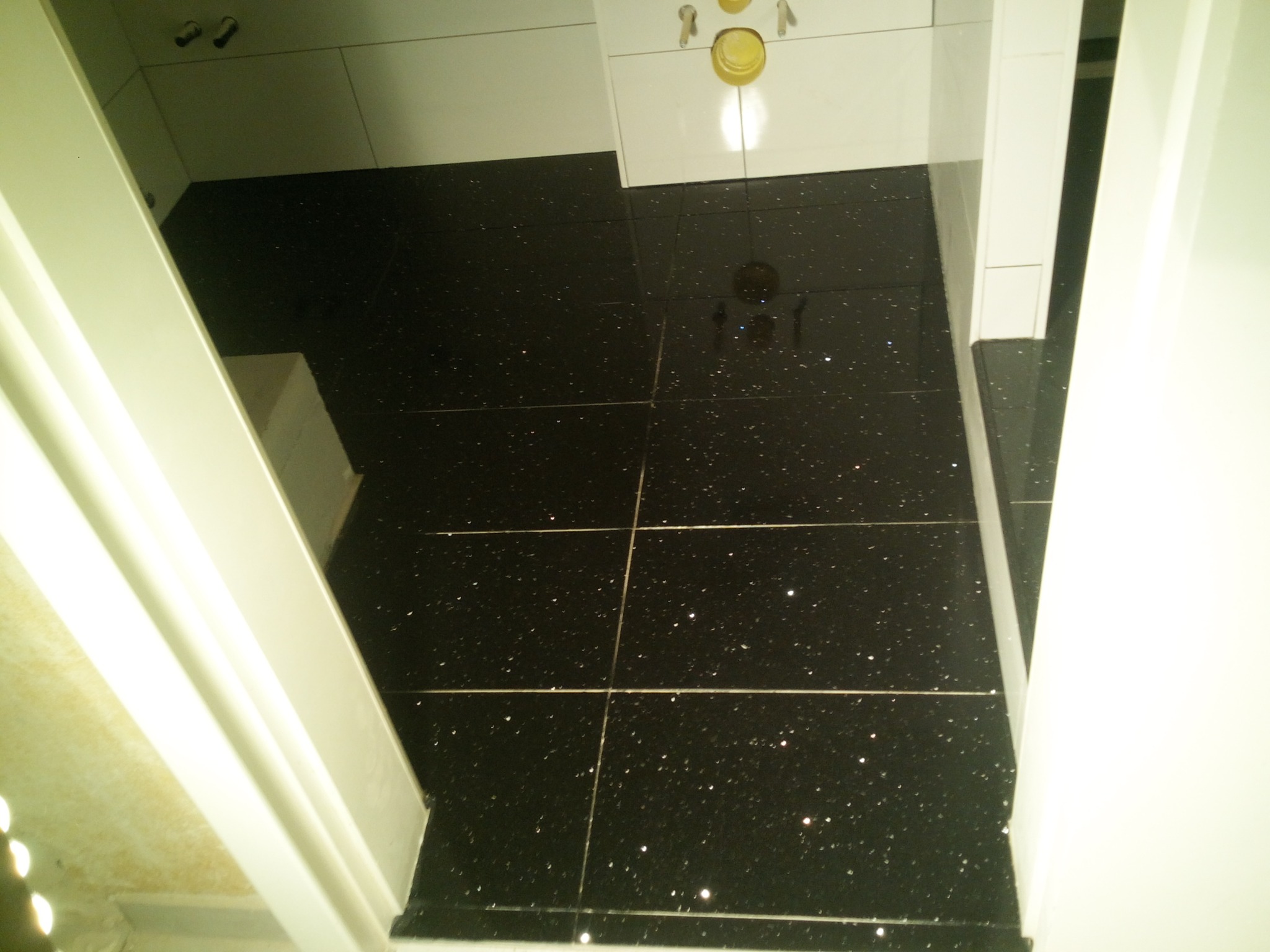 Błyszcząca, czarna podłoga z płytek w łazience, widok z góry, białe wykończenia ścian i drzwi.