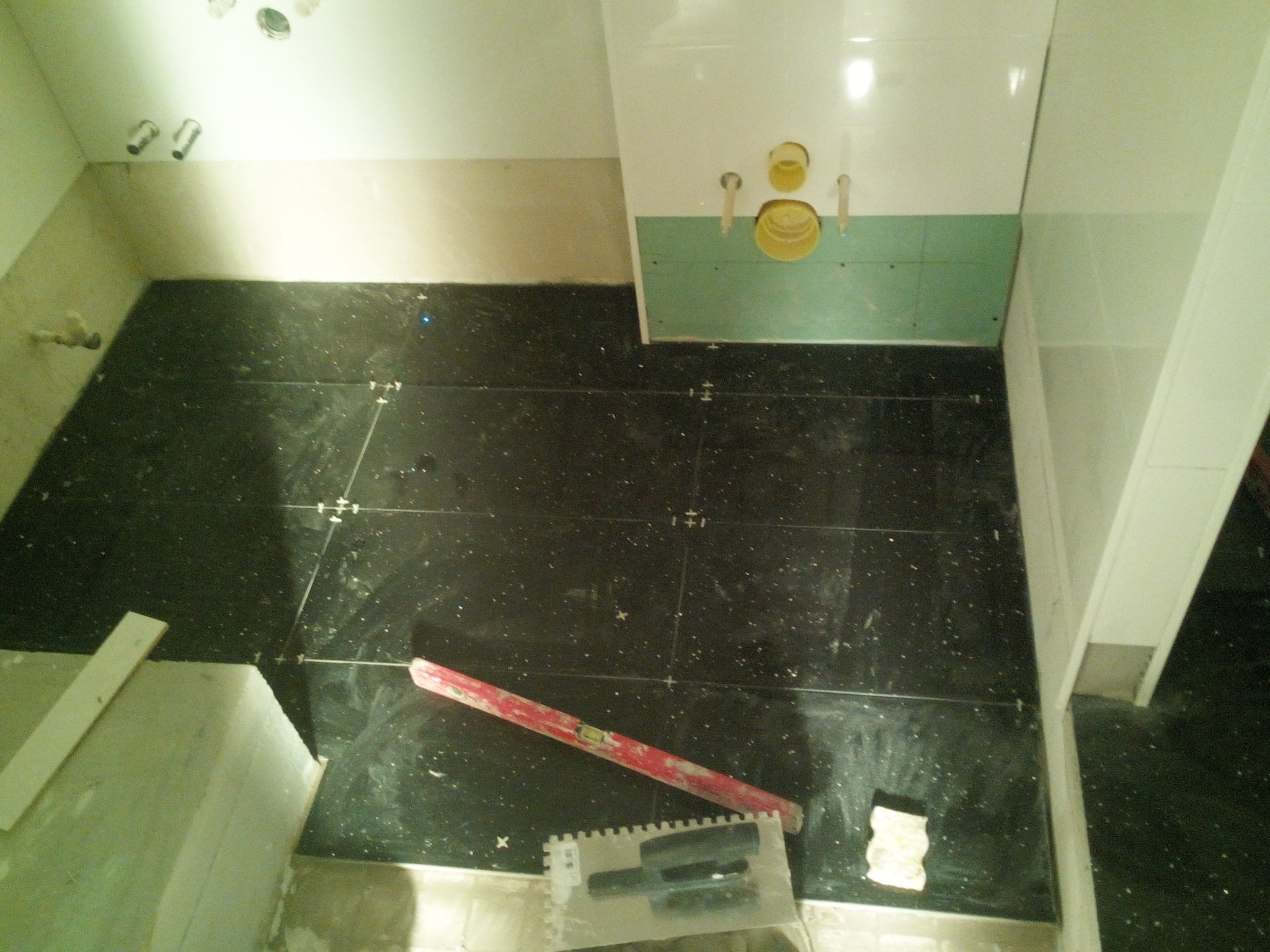 Układanie czarnych płytek podłogowych z białymi drobinkami w łazience, widoczna poziomica, paca do kleju i krzyżyki dystansowe.