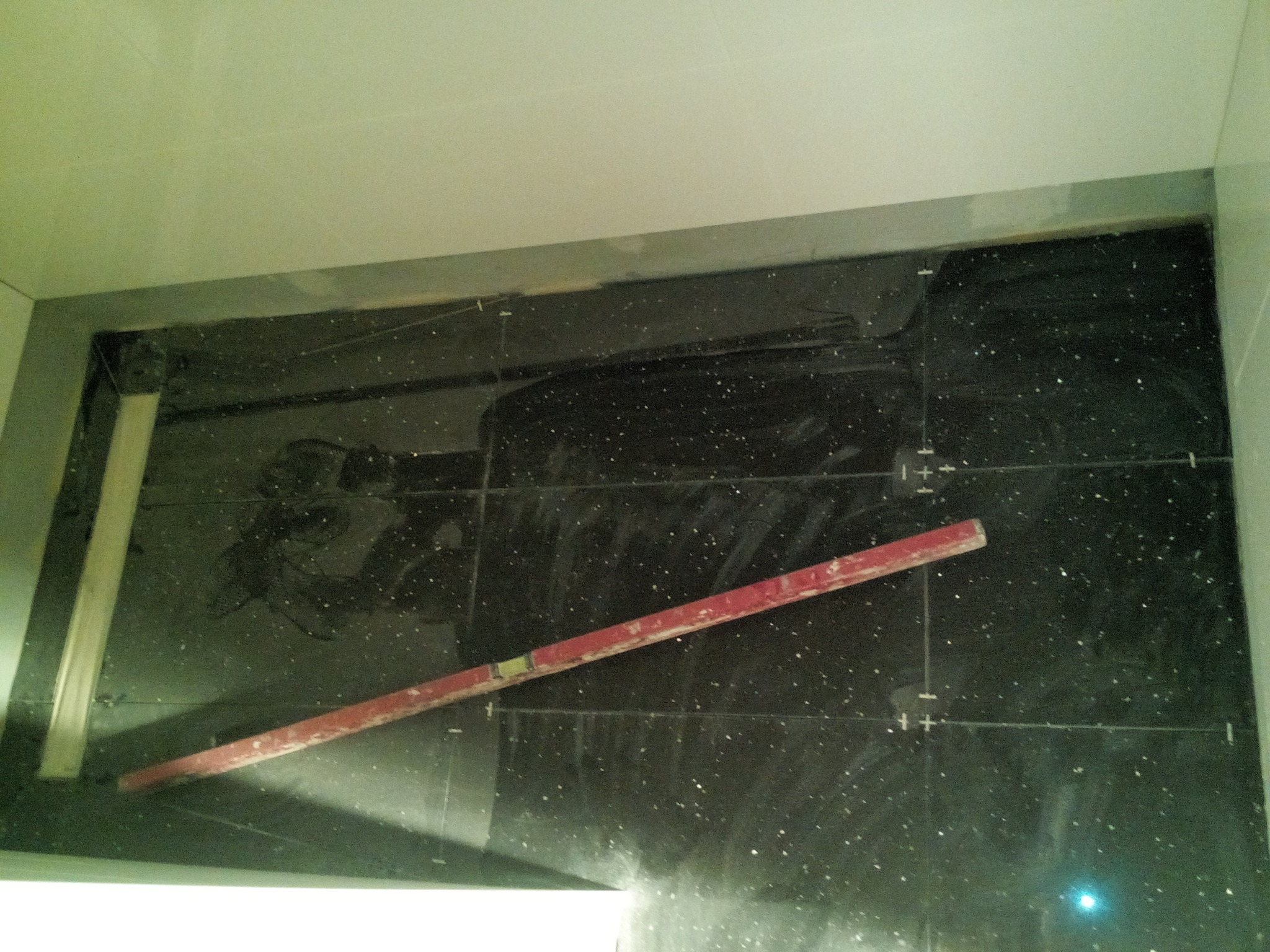 Ciemne, błyszczące płytki ścienne z poziomnicą i listwą, w trakcie układania, widoczne ślady kleju i linie pomocnicze.